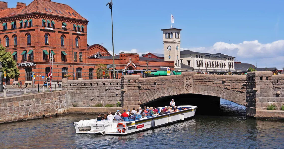 En turistbåd fyldt med passagerer sejler under en stenbro, omgivet af historiske bygninger og et togstationstårn i baggrunden.