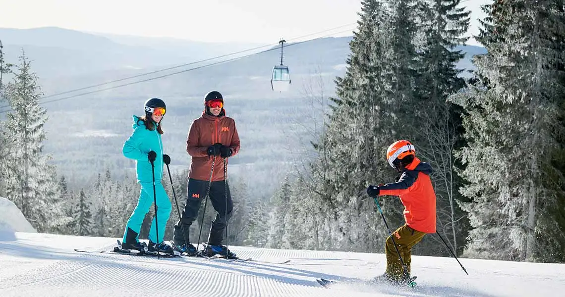 En familie på ski ved Branäs skisted, hvor de står på en velpræpareret piste omgivet af sneklædte træer og bjerge, mens en skilift kører i baggrunden under en lys vinterhimmel.