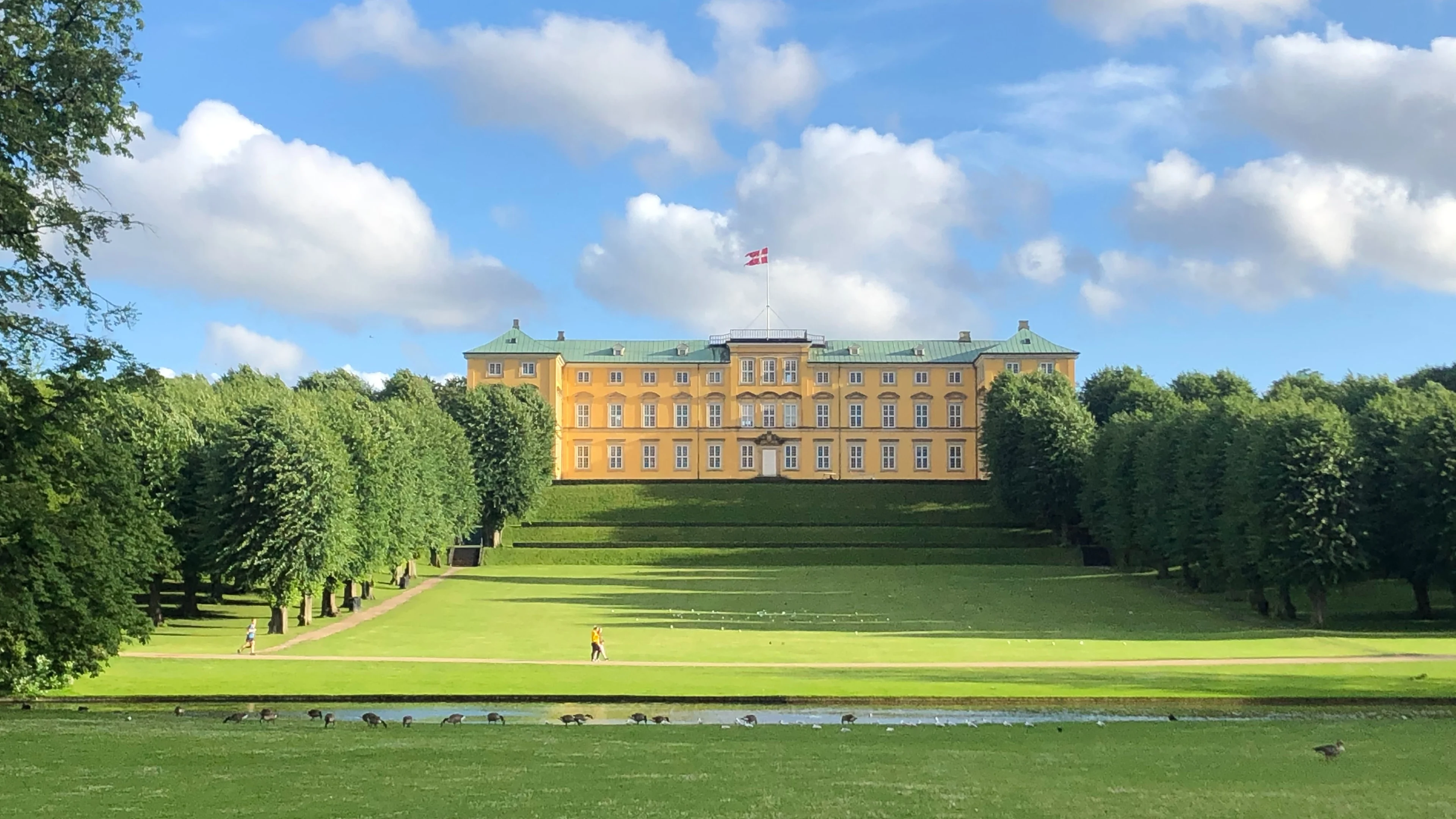 Gult slott omringat av lummiga träd och gräsmattor, med en dansk flagga på toppen av byggnaden under en klarblå himmel.