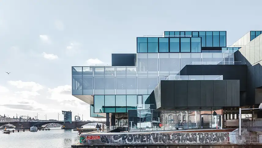 DAC sett utifrån. Byggnaden är byggd i fyrkanter som fogats samman i olika höjder, med glasväggar och ståldetaljer. I bakgrunden syns vattnet i hamnen samt bron Langebro.