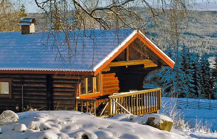 En charmerende skihytte i Sverige, dækket af sne og omgivet af et vinterlandskab med bjerge og skove i baggrunden, badet i sollys på en klar vinterdag.