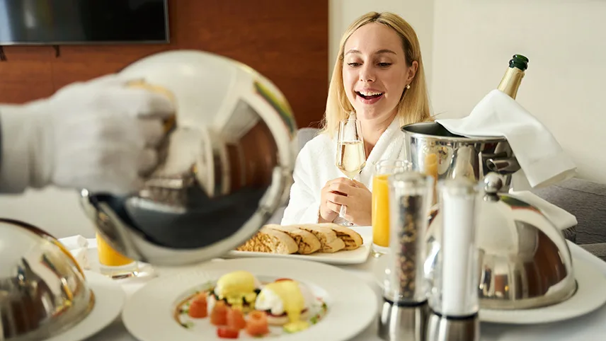En kvinna sitter i en säng på ett hotell och får frukost serverad på en bricka.