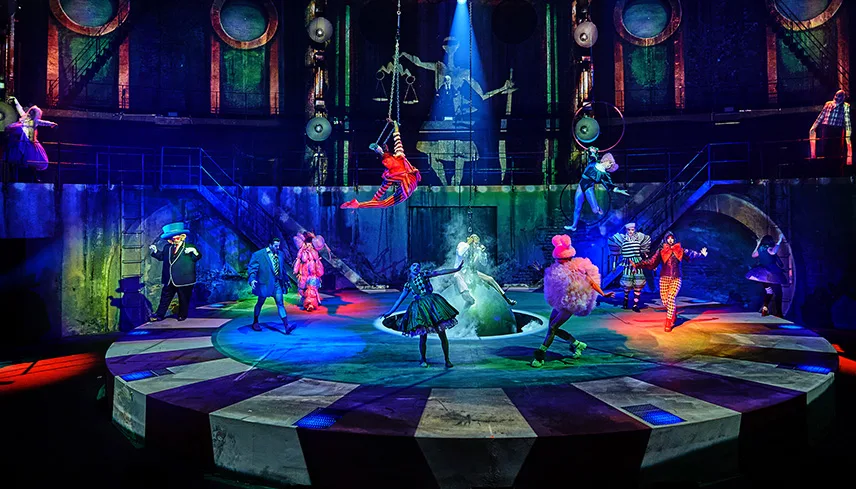 Bild på scenen tagen med lite avstånd. Scenen är rund och belyst med strålkastare i olika färger. Ett antal skådespelare i dramatiska kostymer dansar och sjunger, och en akrobat hänger från taket i en trapets.