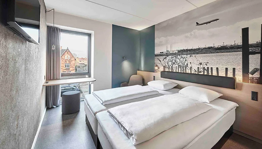 Ett av hotellets dubbelrum med en stor säng bäddad med ljusa sängkläder, tapeter på väggarna och ljusa träslag.