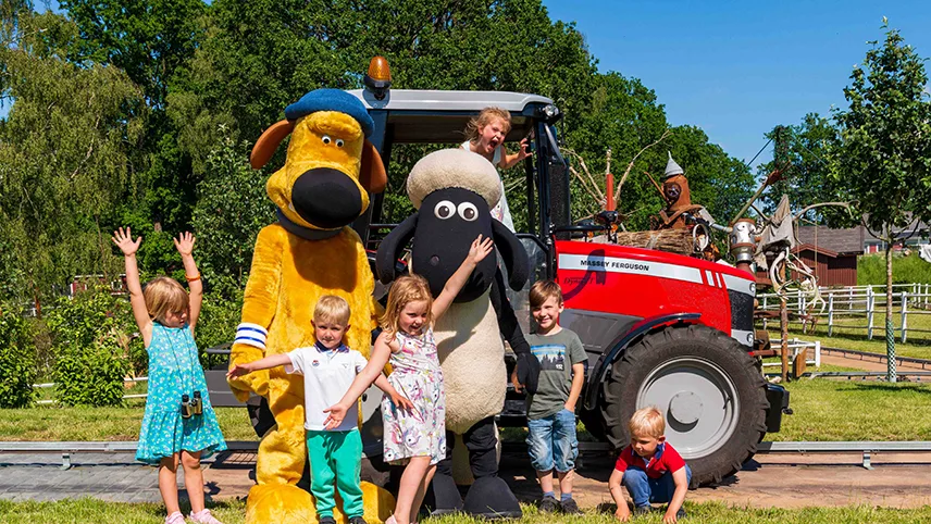 Skånes Djurparks maskot Frode Får omgivet af glade børn en sommerdag. Frode Får står sammen med børnene foran en traktor og sammen med et andet dyr.