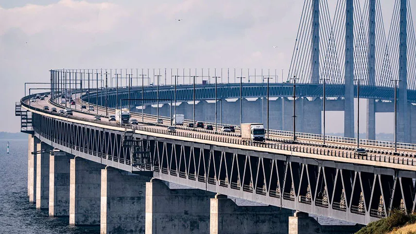 A side view of the Øresund Bridge with cars and trucks driving over and the pylons in the background.