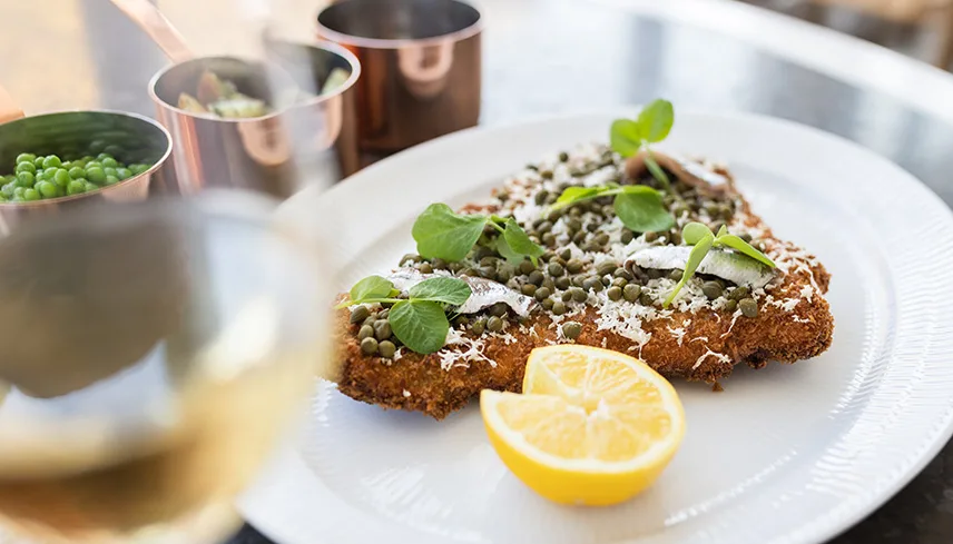 En tallrik med panerad, stekt rödspätta med kapris, pepparrot och en grön kvist, samt en citron.