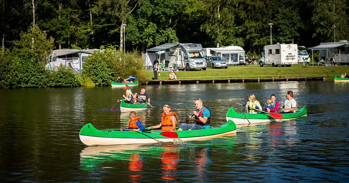 Mennesker padler på en sø med First Camp camping i baggrunden.