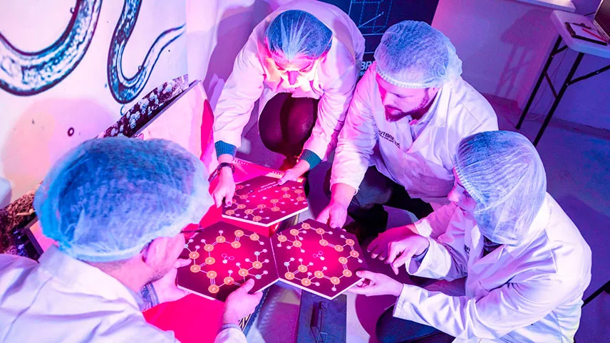 Fyra besökare iklädda vita laboratorierockar och hårnät sitter på golvet och lägger ihop ett stort pussel.