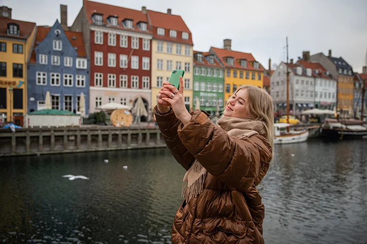 En ung kvinna i dunjacka står vid kanalen i Nyhavn, mot en bakgrund av färgglada hus, och tar en selfie.