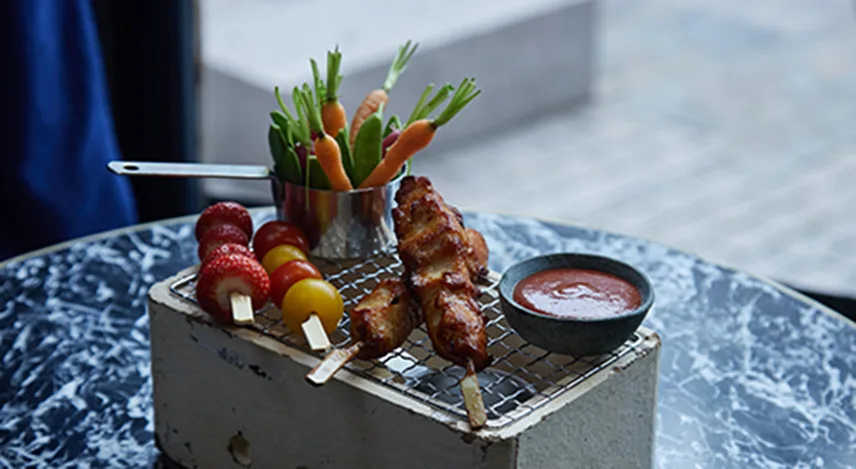 En rätt från Capas barnmeny. Maten är serverad på en speciell tallrik och består av ett kycklingspett, en skål med sås, spett med ugnsbakade tomater och en mugg med morötter och sockerärtor.