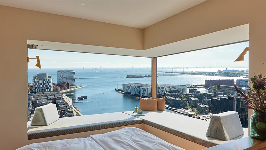 Bild inifrån en av lägenheternas sovrum. Man ser fotändan på en bäddad säng samt ett stort panoramafönster med utsikt över Nordhavn, hamnen och Øresundsbron i bakgrunden.