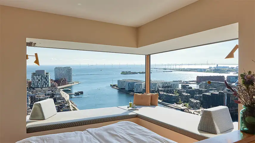 Bild inifrån en av lägenheternas sovrum. Man ser fotändan på en bäddad säng samt ett stort panoramafönster med utsikt över Nordhavn, hamnen och Øresundsbron i bakgrunden.