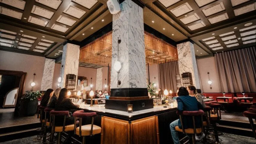 Hotellets bar i form av en fyrkantig bardisk med stora pelare i marmor. En handfull gäster sitter vid bardisken och pratar.