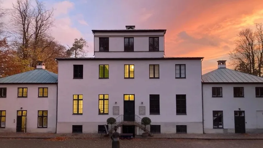 Närbild av huvudbyggnaden på hösten, med färgglada träd och en orange kvällshimmel.