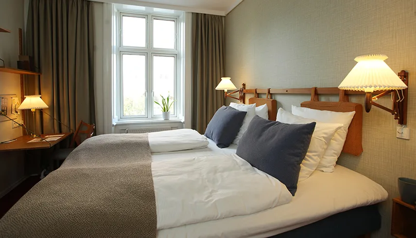 Bild från ett av hotellets dubbelrum i 50-talsstil. Man ser en stor dubbelsäng med vackra textilier och tidstypiska vägglampor ovanför.