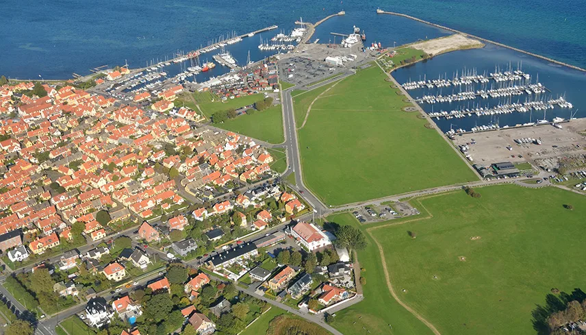 Flybild över orten Dragør. Man ser havet i bakgrunden, hamnen med bryggor och båtar i vattnet, hus och gröna fält.