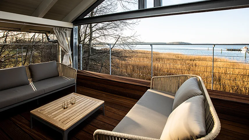En övertäckt veranda till ett av semesterhusen. Man ser två soffor med ett bord och ljuslyktor. I bakgrunden ser man vassen, kala träd och blankt vatten.