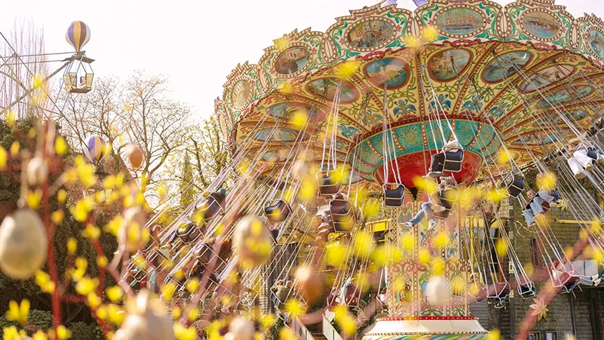 Den klassiska karusellen Slänggungorna är igång och besökare sitter i gungorna och skrattar. I förgrunden syns gula blad och påskägg.