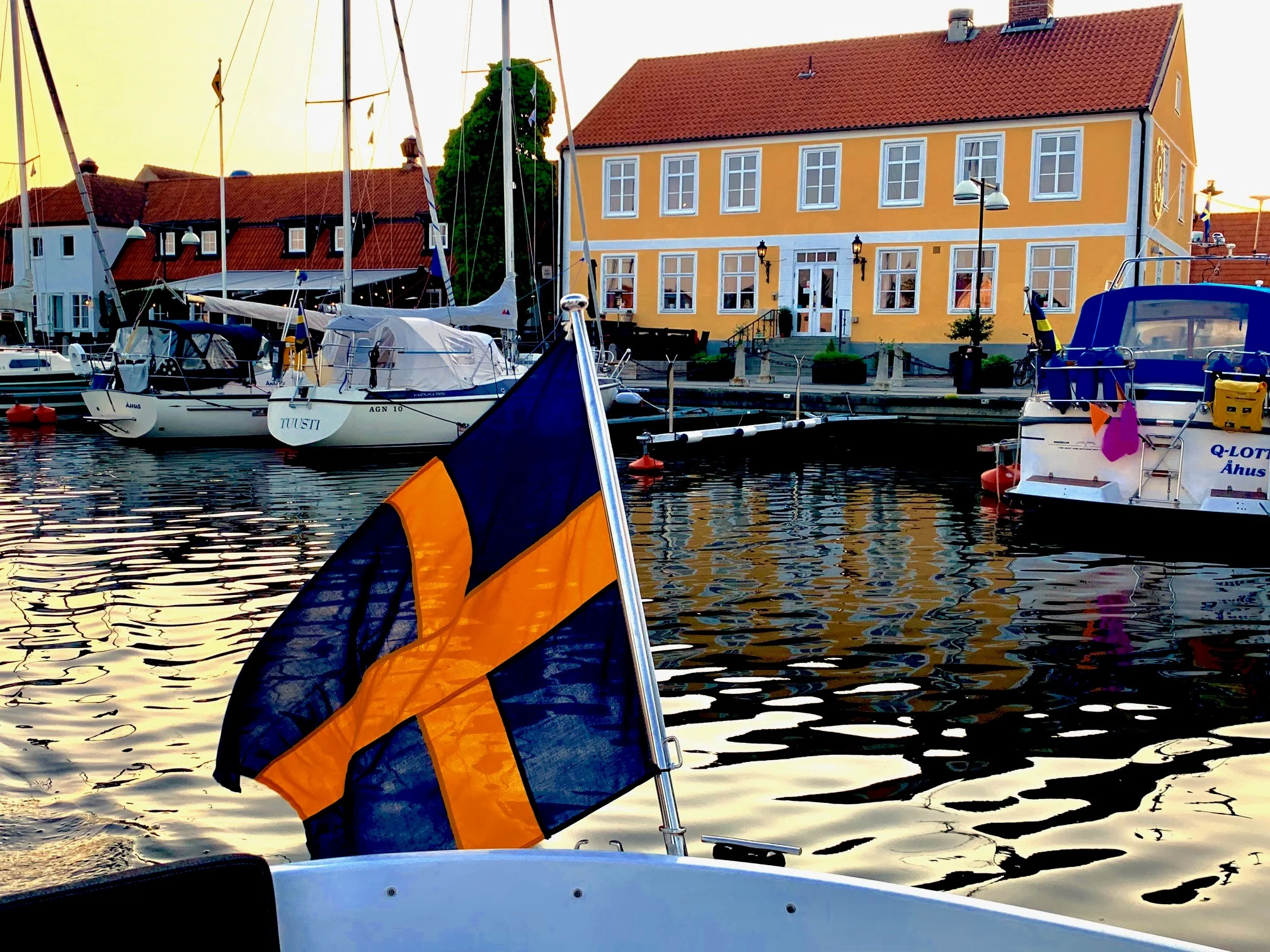 Et svensk flag vajer fra en båd, der ligger ved en kaj, omgivet af andre både og en gul bygning i baggrunden.