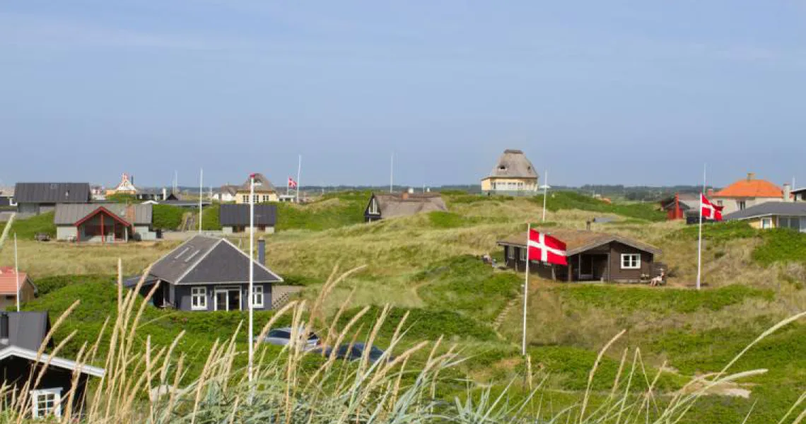 Sommarstugor med danska flaggor som vajar på sanddyner omgivna av gröna kullar under en blå himmel.