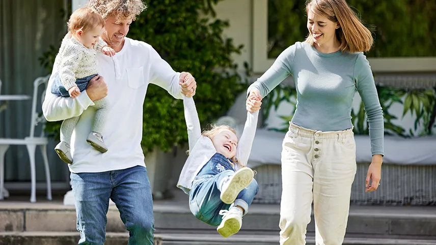 En familj står framför en byggnad. En liten flicka hoppar i luften medan hon håller sin mammas och pappas händer. Pappan har ännu ett barn på ena armen.