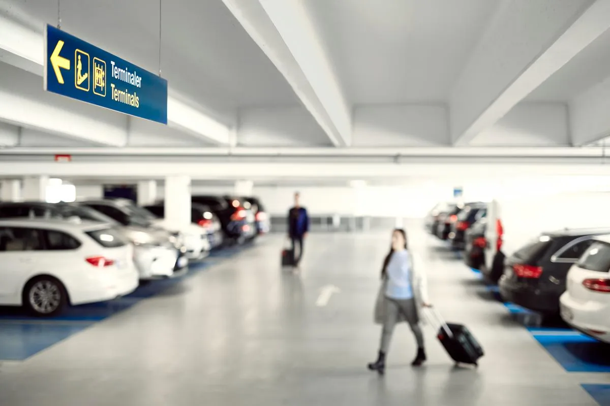 Parkeringsgarage med parkerade bilar och två resenärer som går med varsin resväska.
