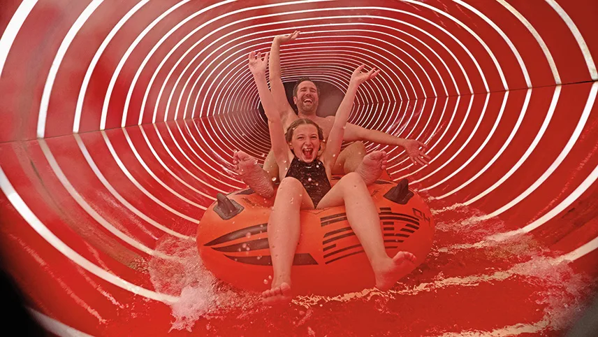Bild från badlandet. Två personer, en vuxen och ett barn, åker i en badring i en vattenrutschkana. Bägge två skrattar och har händerna uppe i luften.