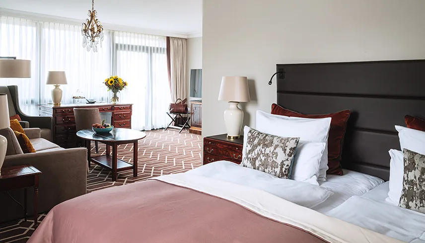 Ett av hotellets sviter. I förgrunden syns en stor dubbelsäng i mörkt trä, bäddad med vita och rosa sänkläder. I bakgrunden ser man stora fönster med skira gardiner, en vacker sittgrupp och en äkta matta på golvet.