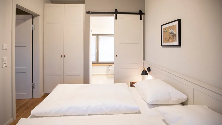 Bild från ett sovrum i ett av gästhusen. Här ser man en bred dubbelsäng bäddad med vita sängkläder, en garderob och ett fönster där solen lyser in.
