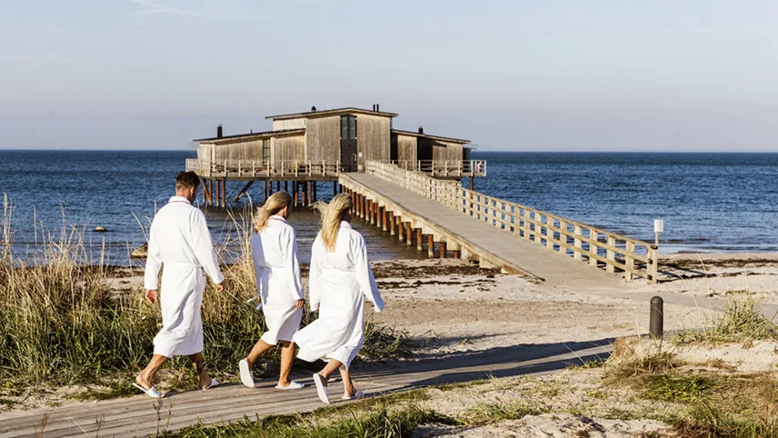 Tre gæster klædt i hvide badekåber på vej mod Koldbadehuset på Hotel Skansen, som ligger ude i havet med adgang fra hotellets badebro.