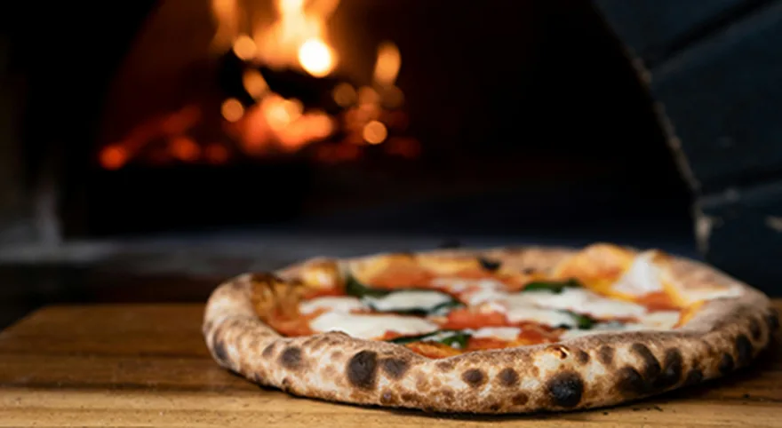 Bild av en pizza med tomatsås, ost och basilika på ett brunt träbord framför en brasa.