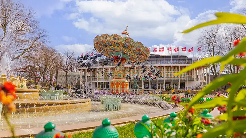 I förgrunden syns en fontän med vatten i tunna strålar, blomstrande planteringar och i bakgrunden ser man en karusell som snurrar.