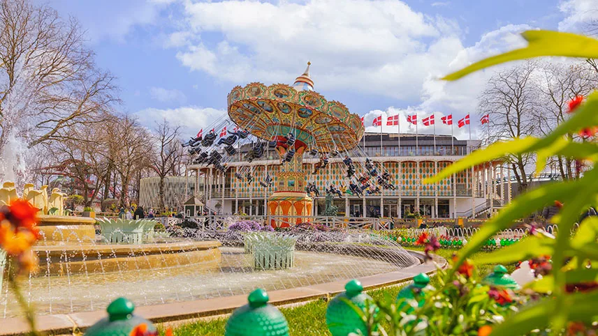 I förgrunden syns en fontän med vatten i tunna strålar, blomstrande planteringar och i bakgrunden ser man en karusell som snurrar.