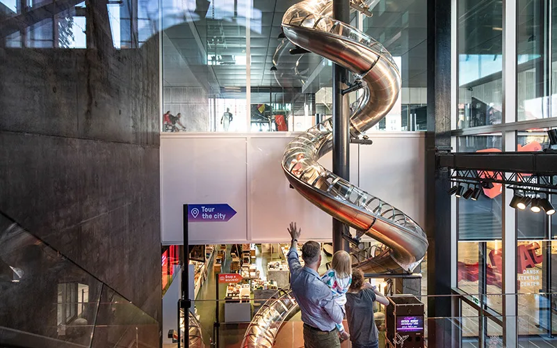 På bilden syns rutschkanan som går genom hela museet. En person står med ryggen mot kameran och håller i ett barn och vinkar till någon som är på väg att sätta sig i rutschkanan.