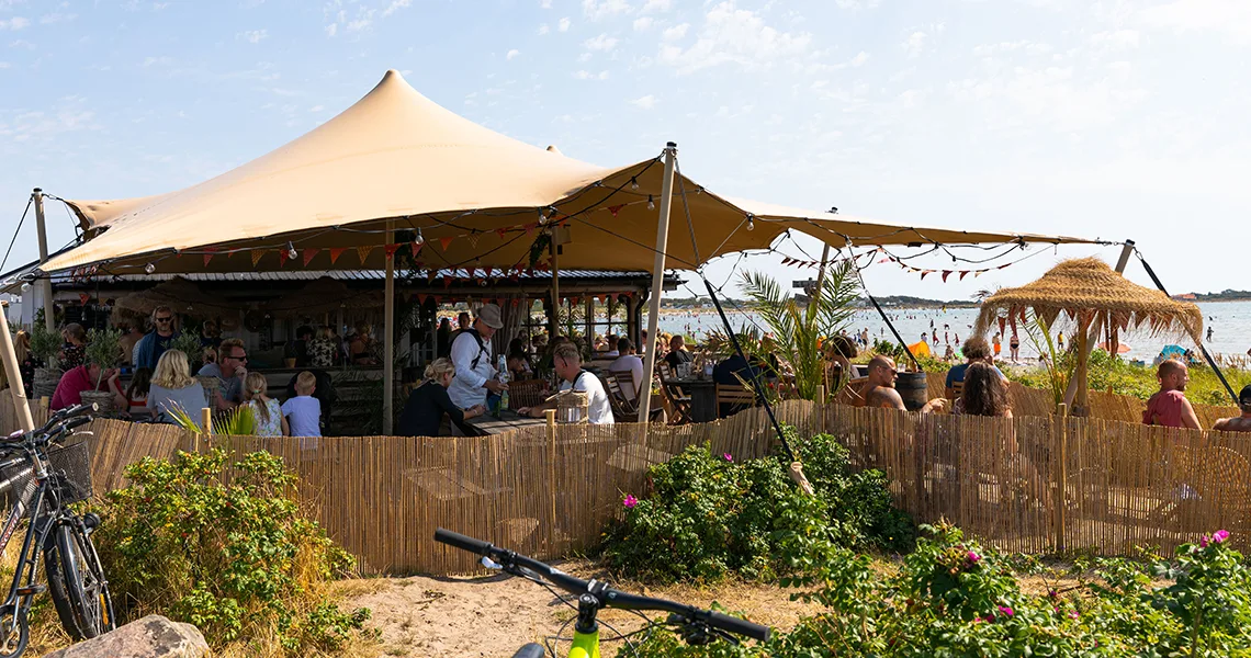Halland Brittas strandveranda hvor mennesker fester under en stor pavillon på stranden.