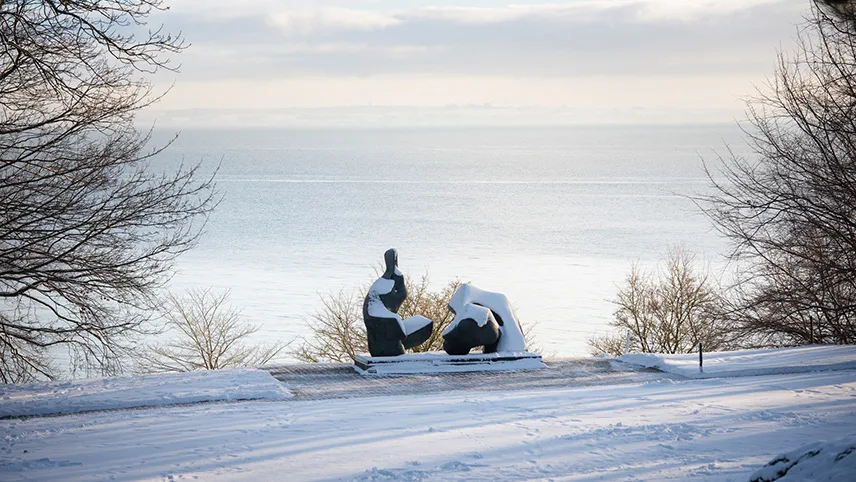 Vinter vid Louisiana - bild tagen från skulpturparken och ut över Öresund. I förgrunden syns snötäckt mark och en staty med ett lager snö. I bakgrunden syns det stilla havet och träd med kala grenar.