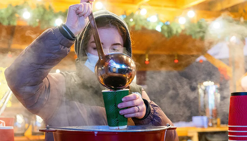 En kvinna häller upp gluhwein i en grön pappmugg med en slev. I bakgrunden syns julpynt och röda lampor.