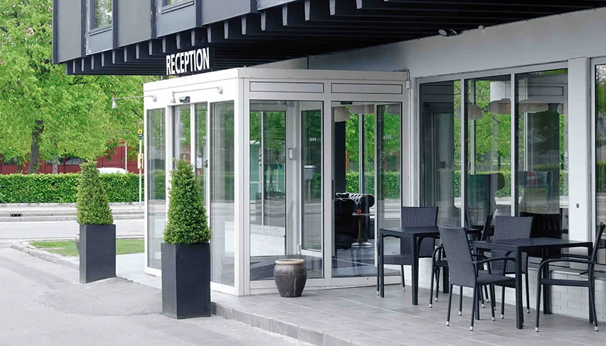 Hotellets fasad med ingång, lummiga träd och utemöbler.