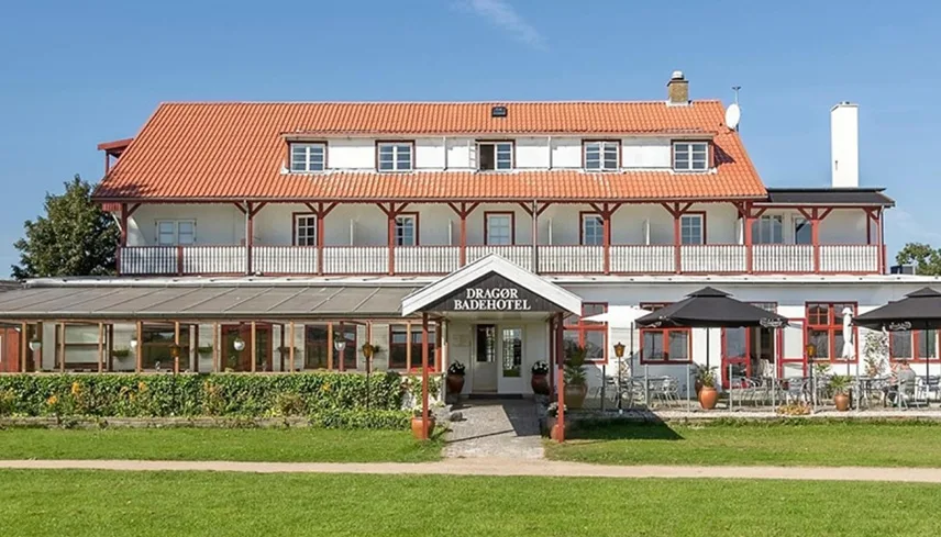 Dragør Badehotel en solig sommardag. Det vita huset med röda detaljer ligger nära vattnet, omgivet av gröna gräsmattor.