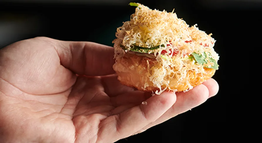 En hand håller i en ugnsbakad krokett som ser ut att innehålla kött, grönsaker och är täckt med riven ost.