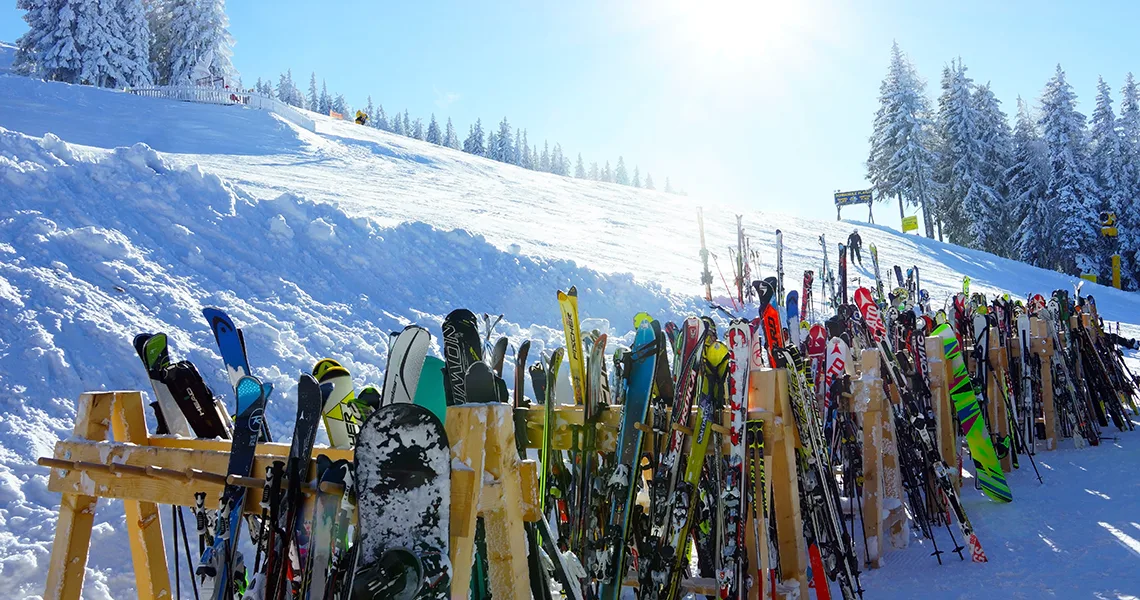 Rækker af ski og snowboards opstillet ved bunden af en sneklædt piste i Sälen, omgivet af skov og skinnende sollys på en klar vinterdag.