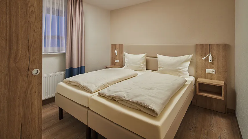 Sovrummet i ett av semesterhusen. En bred dubbelsäng är bäddad med ljusa sängkläder, väggarna är målade i vitt och beige och man ser ett fönster med gardiner.