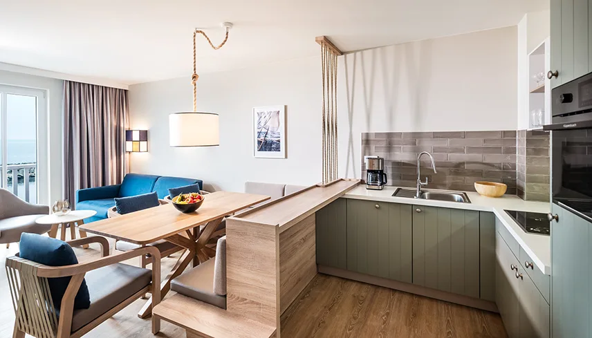 Bild från en av hotellets lägenheter. Till höger syns ett kök med gröna skåpluckor, köksö i ljust trä och en handvask. Till vänster syns köksbord och en soffgrupp.