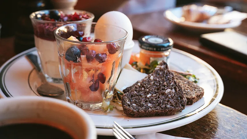 En frukosttallrik från Granola med rågbröd, kokt ägg, marmelad, ett glas med yoghurt och granola och ett glas med fruktsallad.