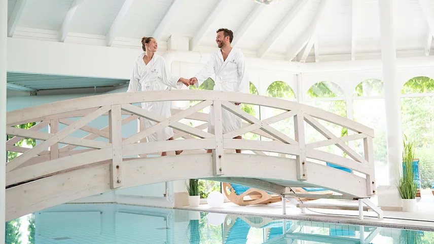 Weissenhäuser Strands spa. Två personer står på en vit träbro som sträcker sig över en pool, de håller varandra i handen och är klädda i vita badrockar. I bakgrunden syns gröna växter och solen faller in genom de stora fönstren.
