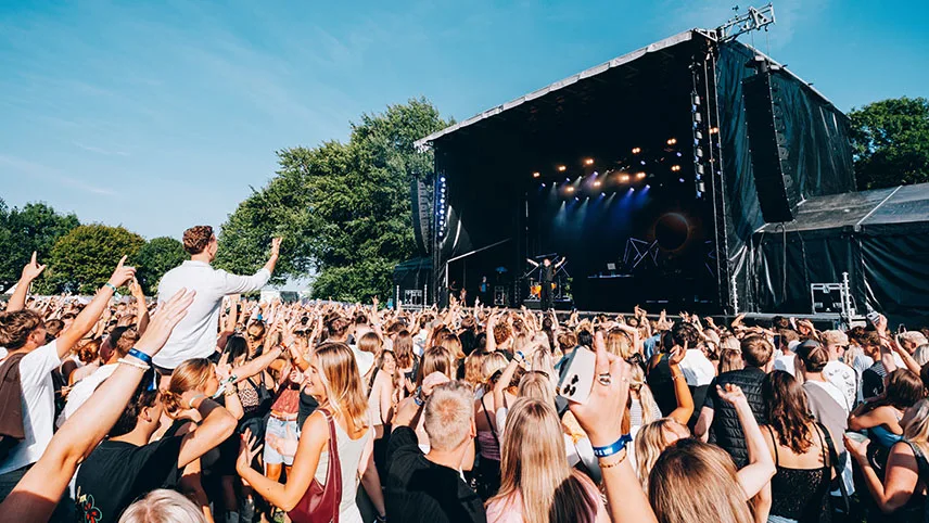 Bild av publikhavet vid förra årets AiaSound. I bakgrunden syns en stor scen mot en blå himmel och gröna träd. Publiken sträcker upp armarna i luften och dansar.