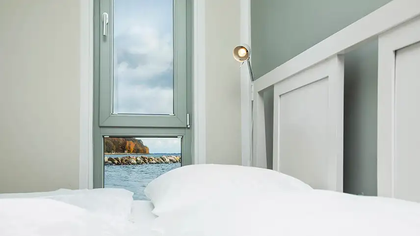 Ett rum sett inifrån med havet, en stenpir och färgsprakande träd utanför fönstret.