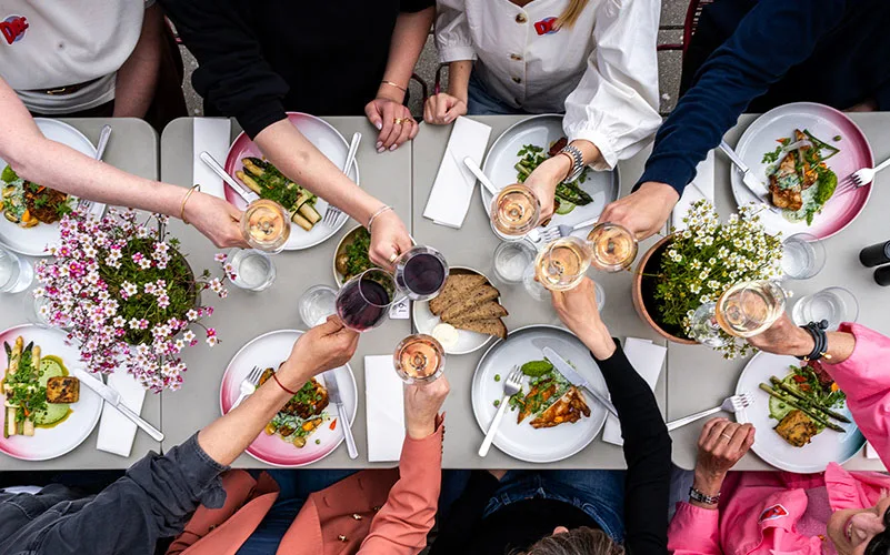 Bild tagen uppifrån av ett långbord i caféet. Bordet är vackert dukat med tallrikar, glas och blommor och man ser matgästernas händer när de skålar med varandra.
