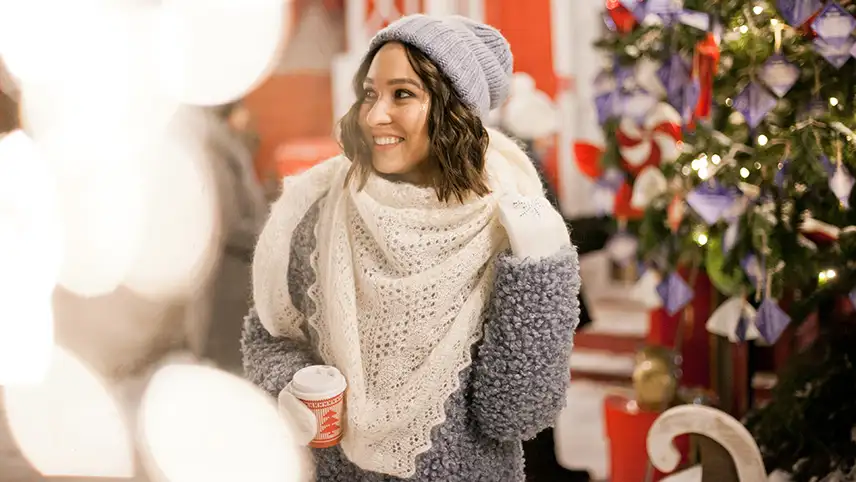 En kvinna iklädd vinterkläder står vid en pyntad julgran och skrattar och dricker något ur en pappmugg med juligt mönster.